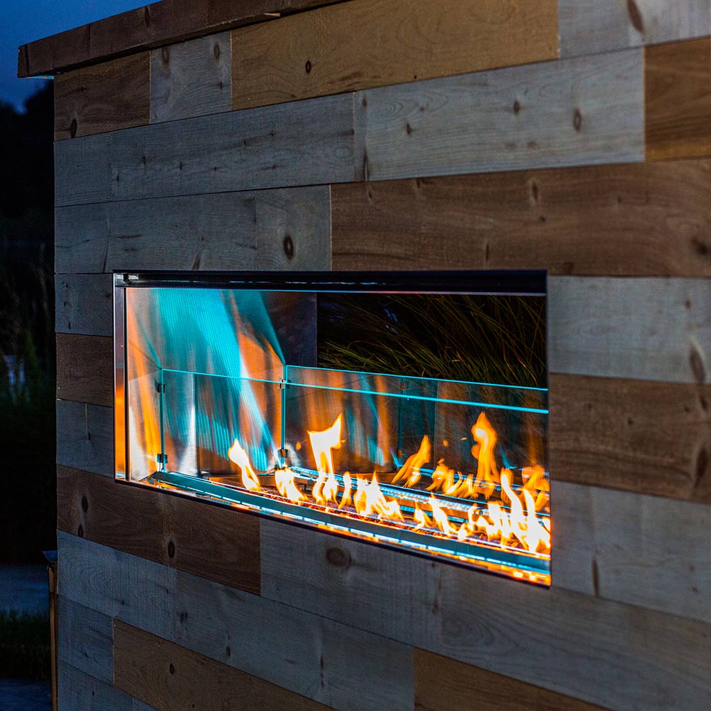 Linear outdoor fireplace insert with glass panel and bright flames behind metal frame